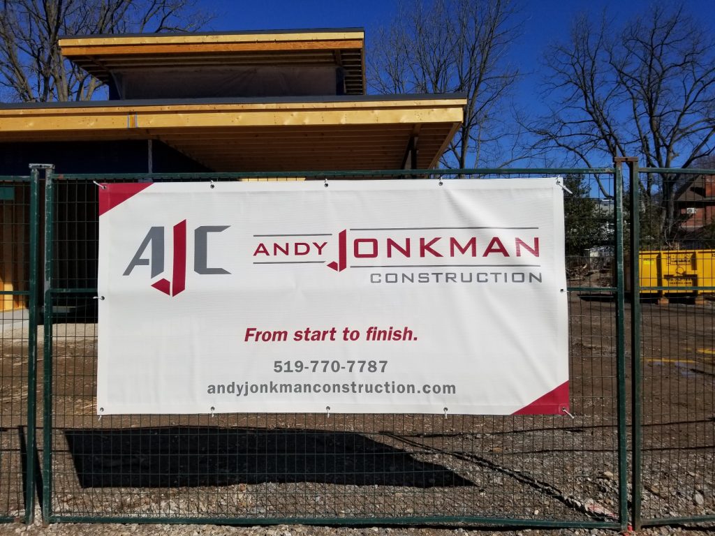 A custom vinyl mesh banner hangs from a temporary fence in front of a house under construction.