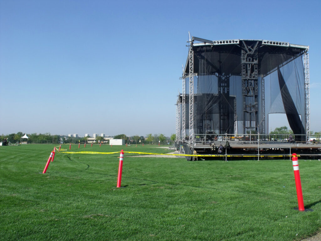 A large stage in the middle of a field. Orange delineators with caution tape encircle the stage.